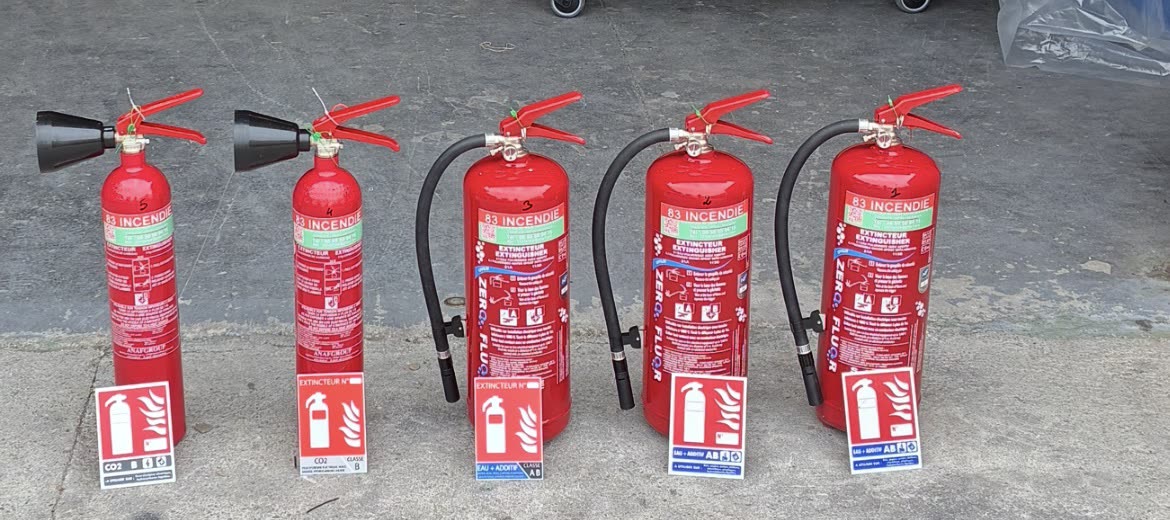 Cinq extincteurs 83 incendie alignés en atelier avec leurs panneaux de signalisation, prêts à être installés